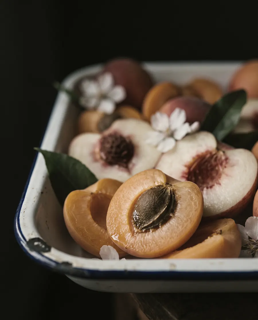 Halved apricots and white peaches in a chilled enamel tray, candlelight raking across the cut fruit.
